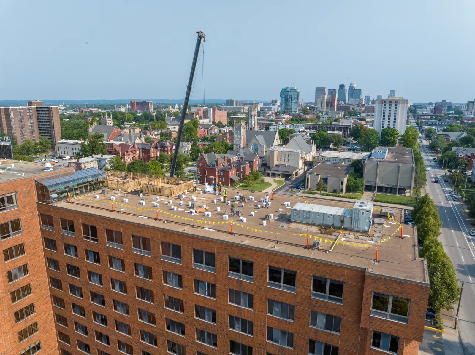 Crew working on the main roof mid-project with a crane boom overhead and downtown Louisville behind